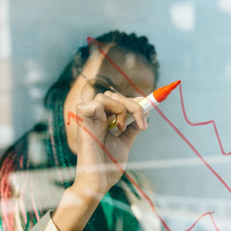 Woman in a Beige Coat Writing on a Glass Panel Using a Whiteboard Marker